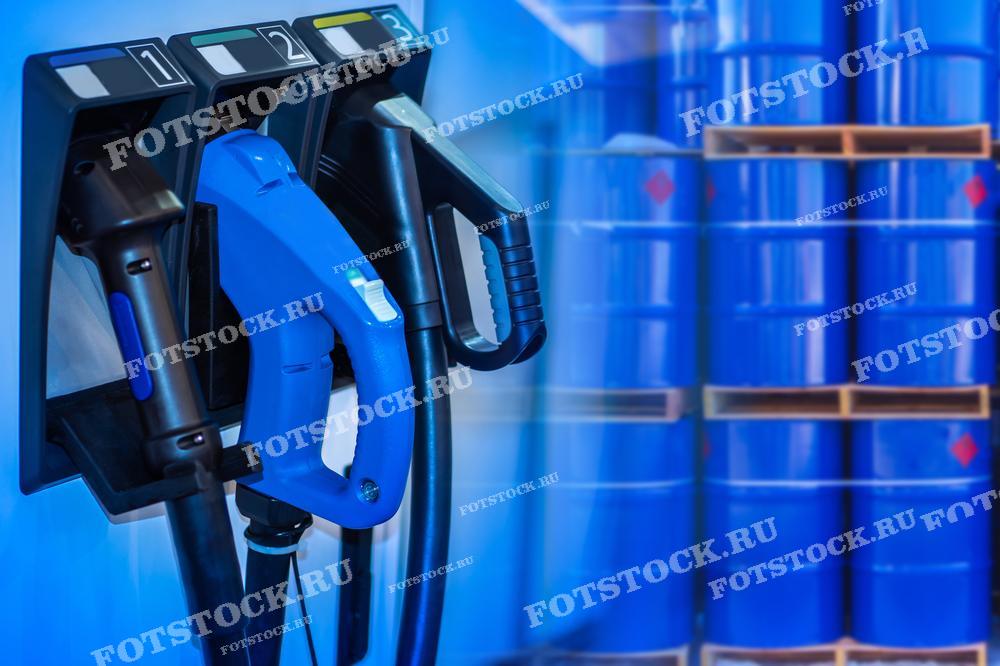 На изображении показаны топливные пистолеты на заправочной станции на фоне синих металлических бочек, используемых для хранения топлива. Это символизирует процесс заправки автомобилей и хранения нефтепродуктов на автозаправочной станции.